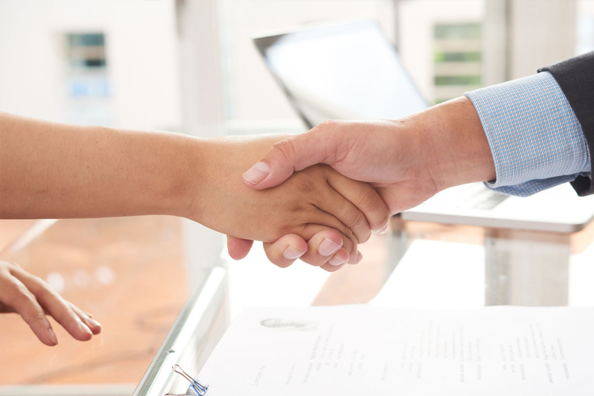 Two people shaking hands across a desk with documents and a laptop visible