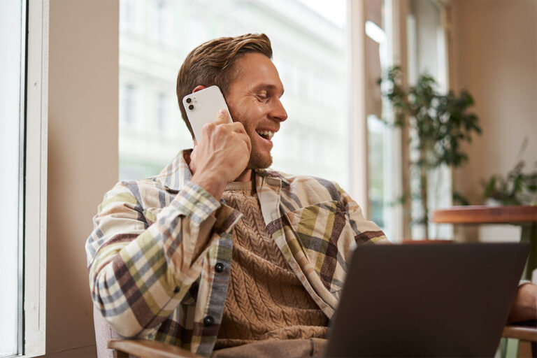 A man smiling while talking on a smartphone and working on a laptop